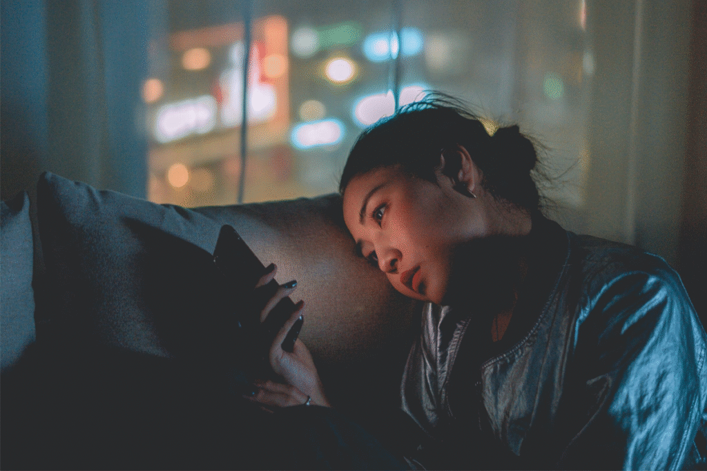Young woman sitting in the dark, illuminated by her smartphone screen, staring thoughtfully — symbolizing emotional connection with technology.