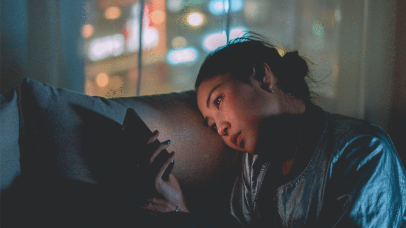 Young woman sitting in the dark, illuminated by her smartphone screen, staring thoughtfully — symbolizing emotional connection with technology.