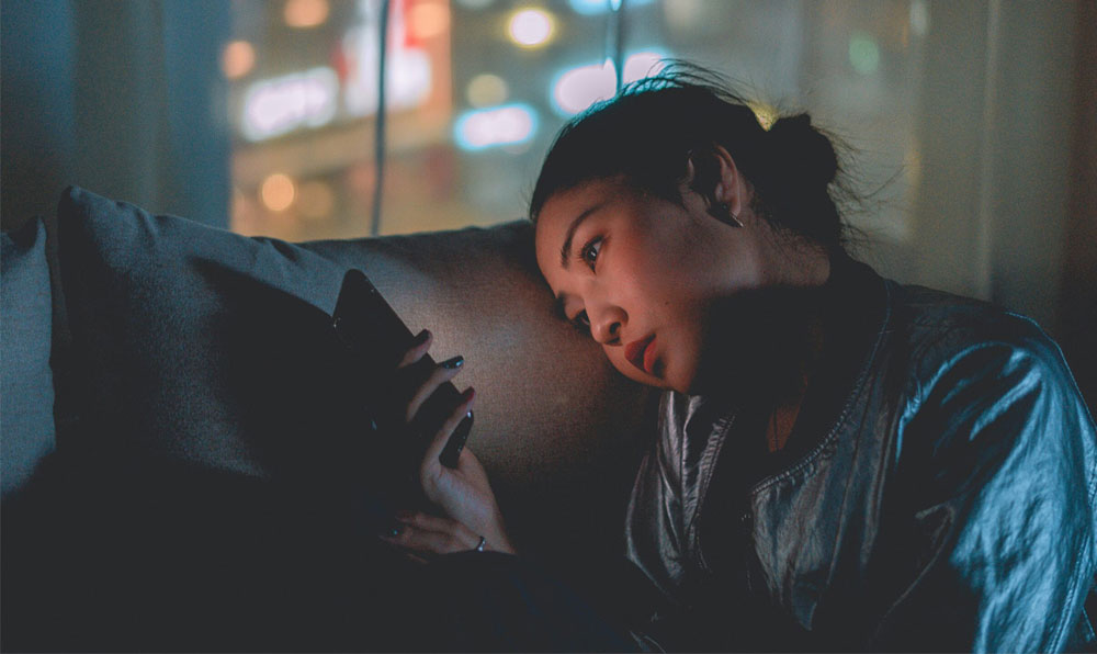 Young woman sitting in the dark, illuminated by her smartphone screen, staring thoughtfully — symbolizing emotional connection with technology.