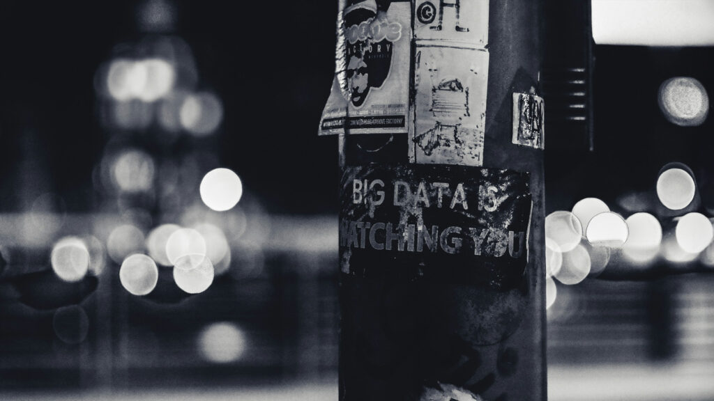 A black-and-white photo of a lamppost covered in worn stickers, one of which reads “BIG DATA IS WATCHING YOU.” The blurred city lights in the background create a dystopian feel, underscoring the urgent need to reclaim digital sovereignty.