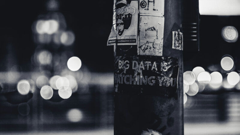 A black-and-white photo of a lamppost covered in worn stickers, one of which reads “BIG DATA IS WATCHING YOU.” The blurred city lights in the background create a dystopian feel, underscoring the urgent need to reclaim digital sovereignty.