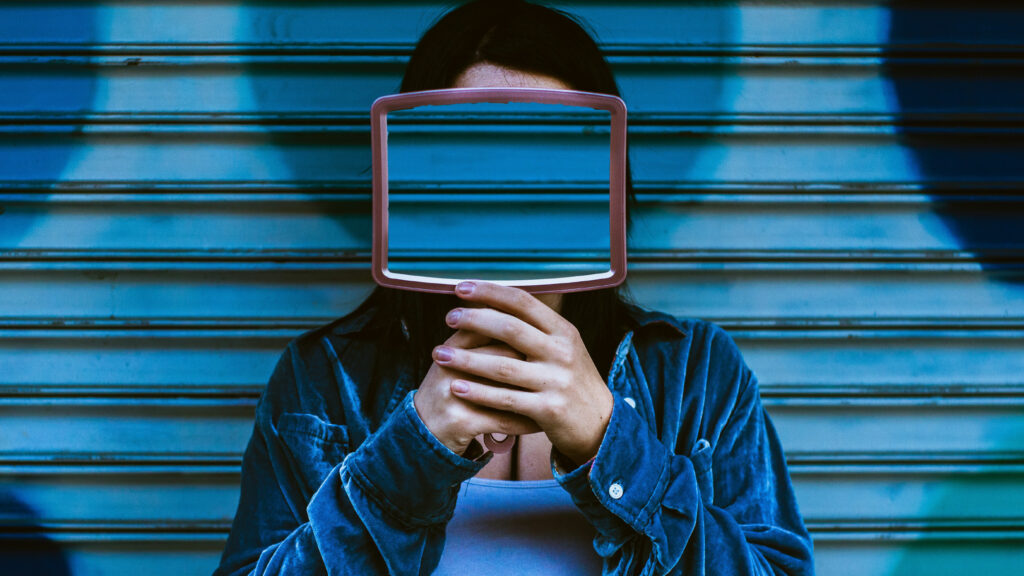 Person holding up a rectangular mirror that replaces their face, reflecting only blue distortion. The background shows a metallic shutter in digital blue tones. A metaphor for emotional loss of self in the age of social media.
