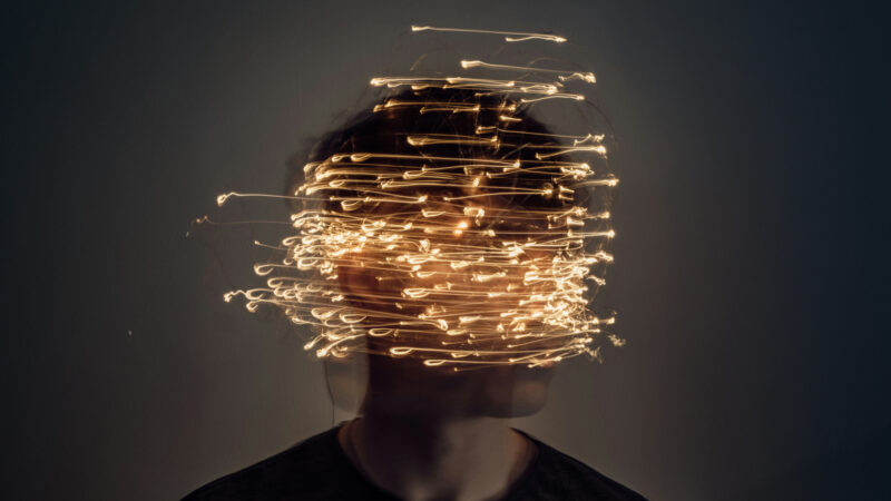 Blurred human face partially obscured by strands of glowing lights, symbolising cognitive overload, digital entanglement, and the loss of mental clarity in the age of algorithmic influence.