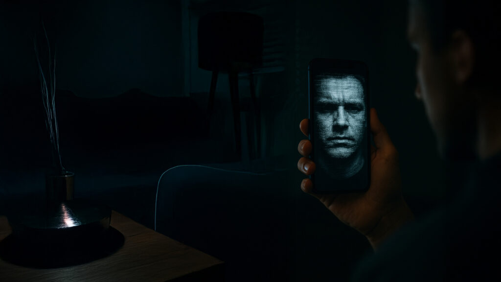 A man holds a smartphone in a dark living room, the screen showing a glitched deepfake face — a visual metaphor for AI in organized crime and the growing threat of digital impersonation.