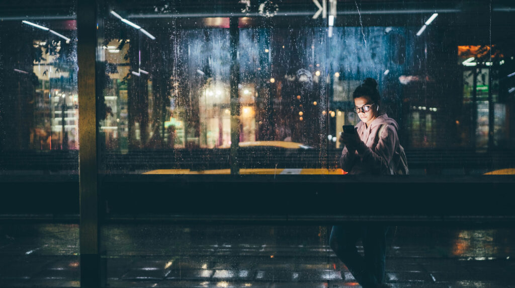 A young woman stands at a bus stop at night, looking at her smartphone through a rain-covered glass wall. City lights reflect in the wet surface, suggesting a quiet moment of connection with conversational AI in an urban setting.