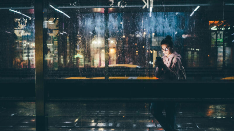 A young woman stands at a bus stop at night, looking at her smartphone through a rain-covered glass wall. City lights reflect in the wet surface, suggesting a quiet moment of connection with conversational AI in an urban setting.