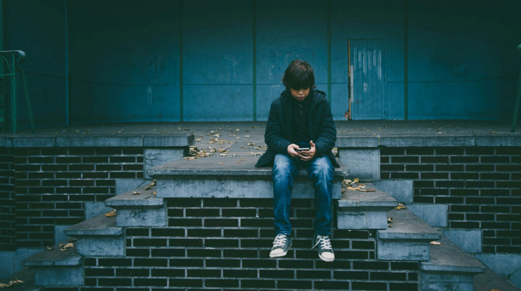 Child sitting alone on steps looking at smartphone, illustrating the social impact of conversational AI and digital loneliness
