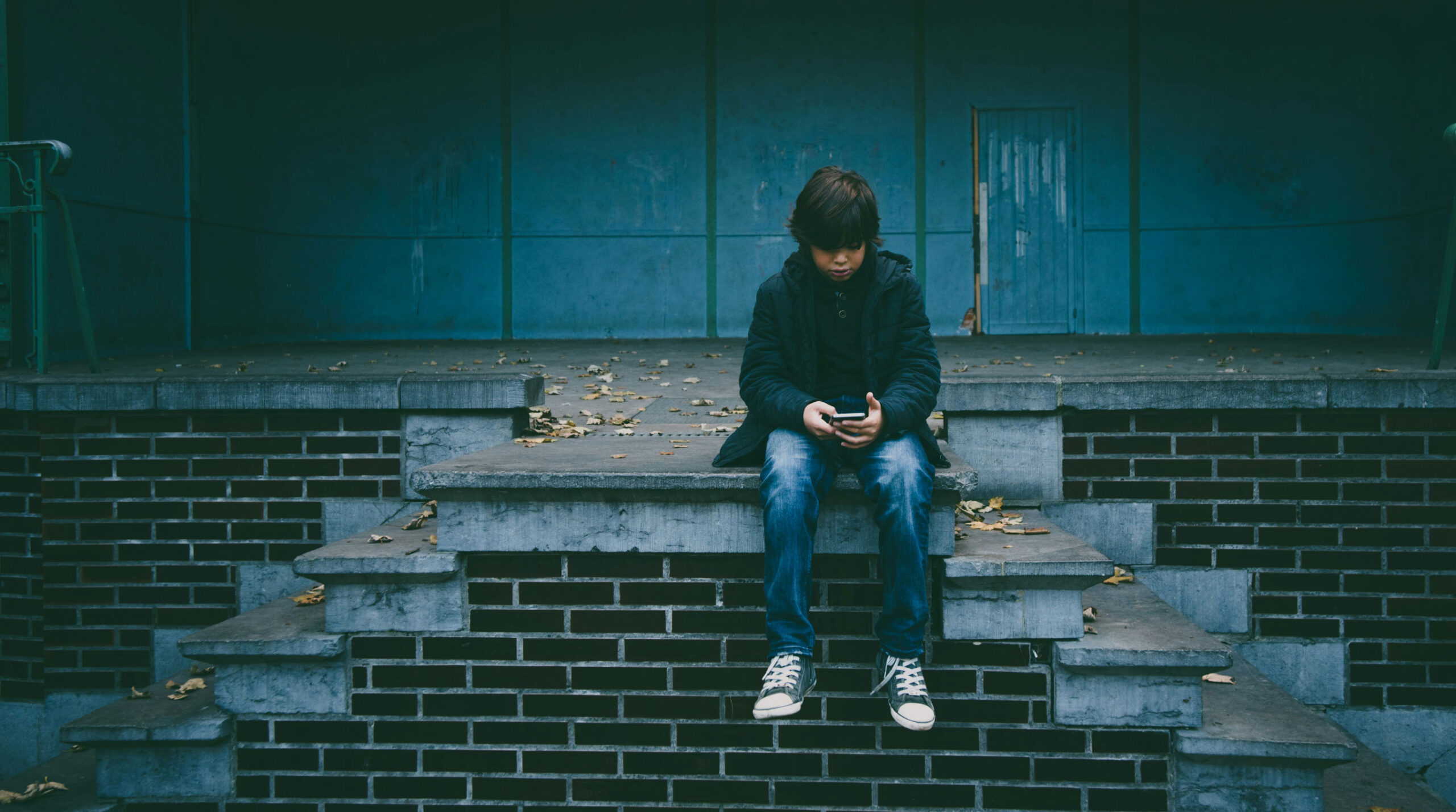 Child sitting alone on steps looking at smartphone, illustrating the social impact of conversational AI and digital loneliness