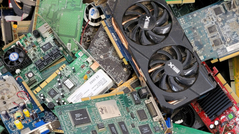Close-up of E-waste showing a large pile of obsolete computer parts, including graphics cards, motherboards, circuit boards, and electronic components awaiting recycling and responsible disposal.
